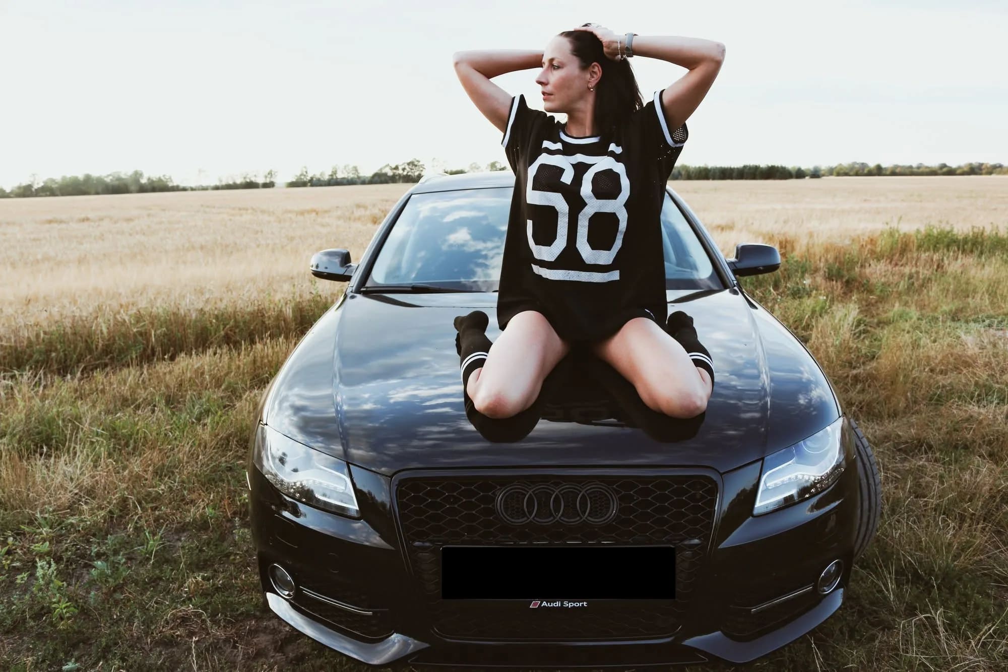Portraitshooting mit Auto: Frau kniet auf der Motorhaube eines schwarzen Audi am Feldrand