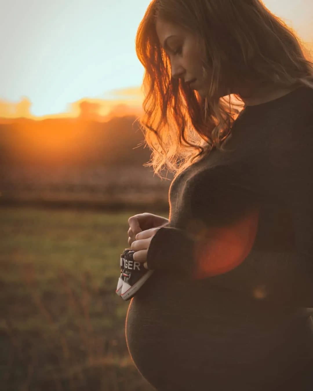 Babybauch-Shooting bei Sonnenuntergang: Schwangere hält kleine Babyschuhe vor den Bauch